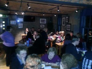 An action shot of the room where the CILIPS Tayside Pub Quiz was held. The quiz master is standing to the left of the picture, overseeing several tables with people gathered around them looking deep in conversation as they work out the correct answers.