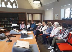 In the same room at the St Andrews University Library Special Collections Unit, the visiting librarians sit on a row of red chairs chatting, in the foreground there is a table with four rare books displayed on cushions.