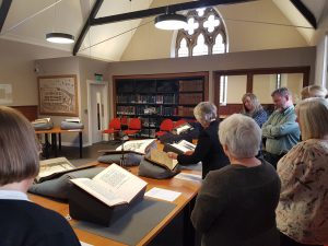 Inside the St Andrews University Library Special Collections Unit, a member of staff looks through a rare book and talks about it to a group of visiting librarians, six of whom are picture gathered around her. There are also four other rare books displayed on cushions on the table in front of the staff member.