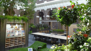a courtyard-style library space with tables, a magazine and book stand, and many hedges, plants and flowers.