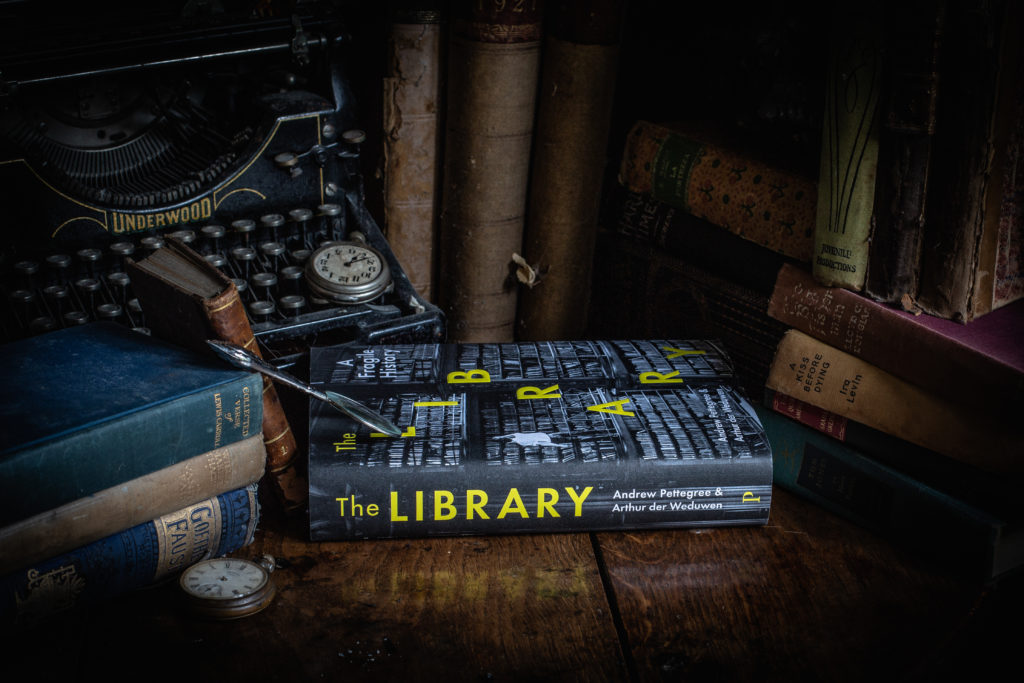 a photograph of The Library: A Fragile History by Andrew Pettegree and Arthur der Weduwen, showing a hardback book with a black/grey library cover photo and yellow text on a wood table with other books and a typewriter nearby