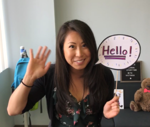 A photograph of Victoria during a library Virtual Storytime, waving and holding a carboard speech bubble that says 'Hello!'