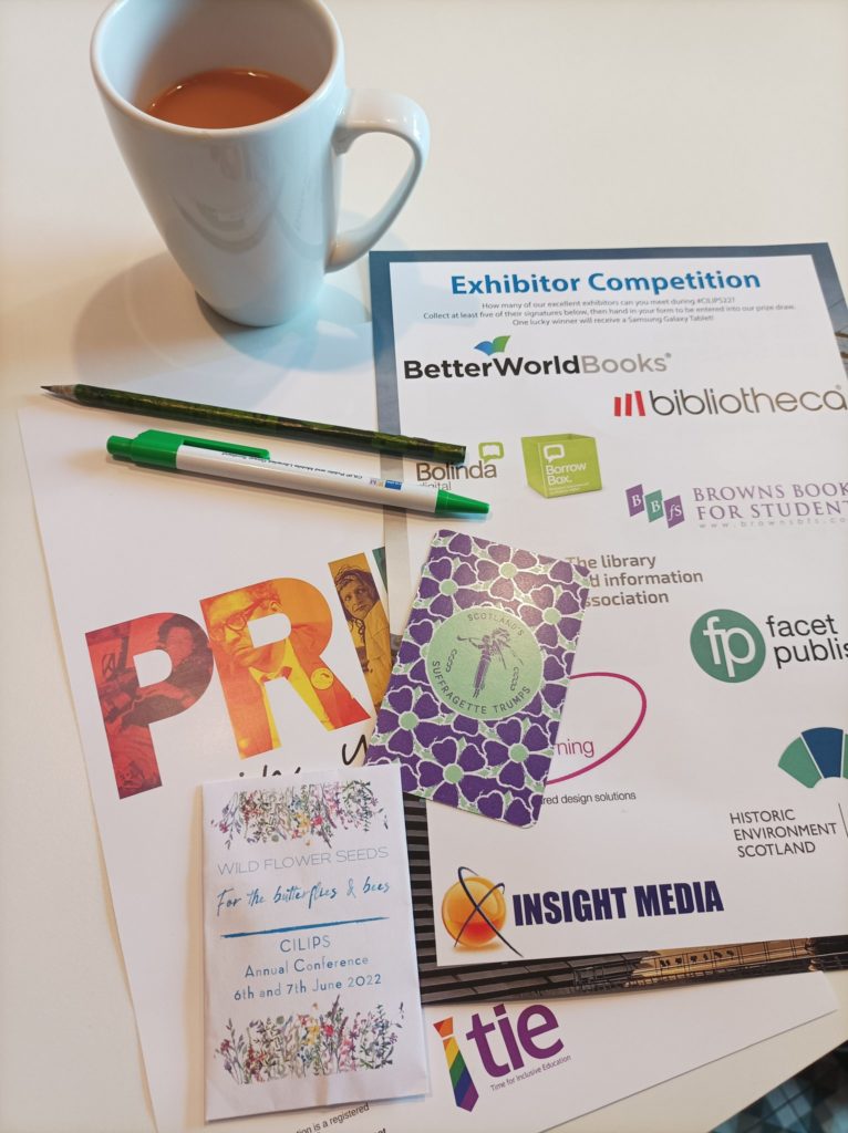 A photograph of the contents of a delegate's conference bag, including wildflower library garden seeds, a newspaper eco-pencil, a TIE Pride poster and a Scottish Suffragette Trump card.