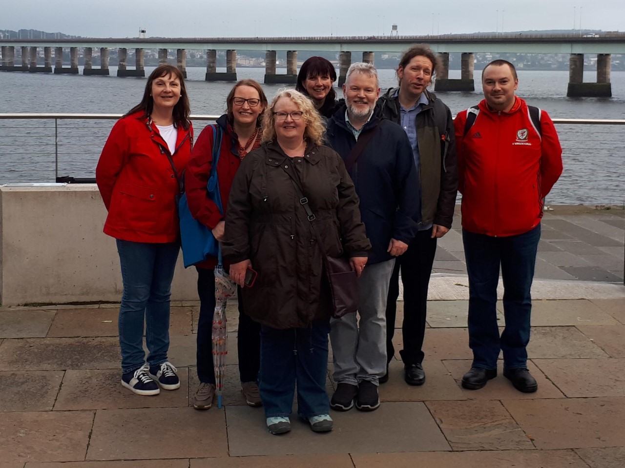 Group of seven people looking towards the camera, the River Tay and concrete Tay Road Bridge are in the background. 