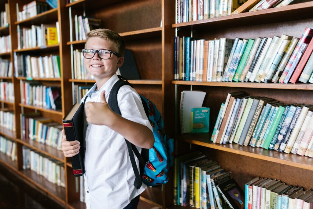 A schoolboy smiling and giving thumbs up in front of school library shelves.