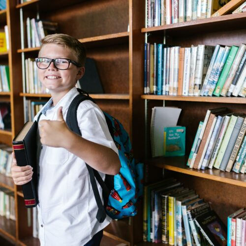 A schoolboy smiling and giving thumbs up in front of school library shelves.