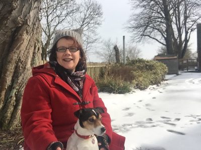 Author sitting with her dog, dressed in a red coat and hat in a snowy landscape.
