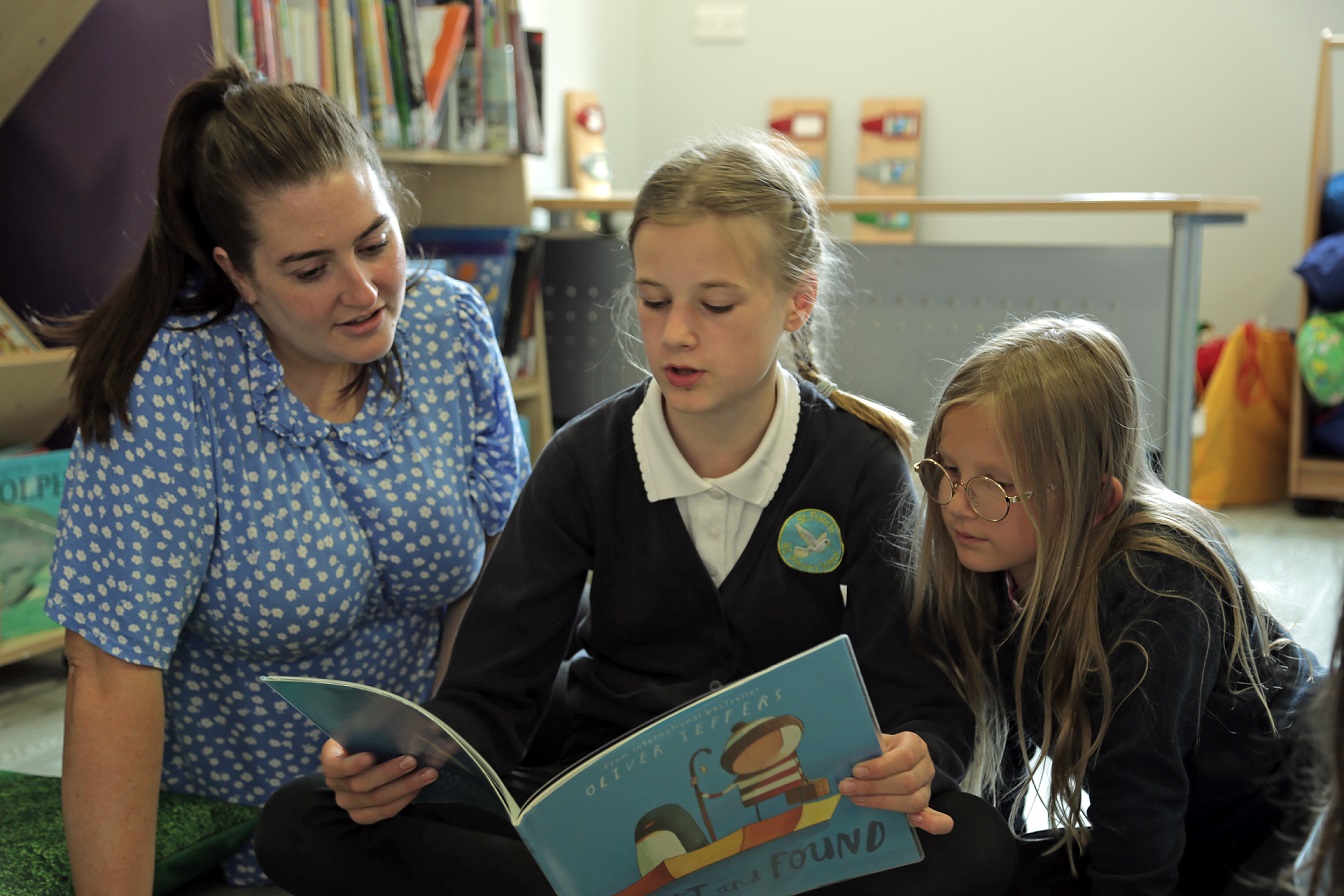 Photograph of teacher sitting to the left of 2 young children whilst they all read a story book.