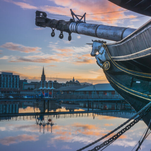 Image of Dundee from the perspective of standing next to the tall ship. Image of Dundee skyline reflected in the water with blue dusk sky and orange clouds.