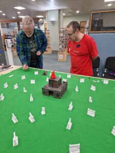 Two people stand over the table top game, action shot where one player is preparing to roll a dice as the other player watches