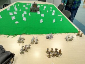 Table top game setup is shown with groups of characters positioned ready to be picked by players. In the background is a green cloth to simulate grass, on this cloth is a series of walls positioned to provide players a strategic hiding place and in the middle is a model building with Star Wars Darth Vader positioned on top.