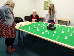 Table top game in action, two people stand and one sits around the table top game which includes in the set a green cloth as the base material with walls, characters, dice and a model building