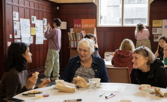 People sitting round a table sharing tea and scones, with people in the backdrop putting up posters on the walls.