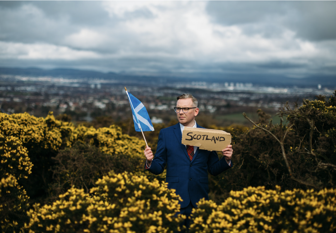 Image of performer Kevin P Gilday in a field with yellow flowers and holding a scotland flag.